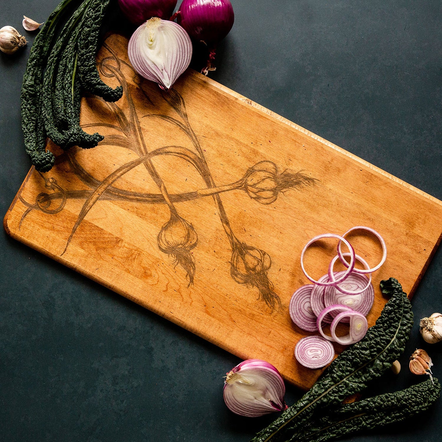 Wooden cutting board with engraved onions surrounded by sliced shallots and whole shallots on a dark surface