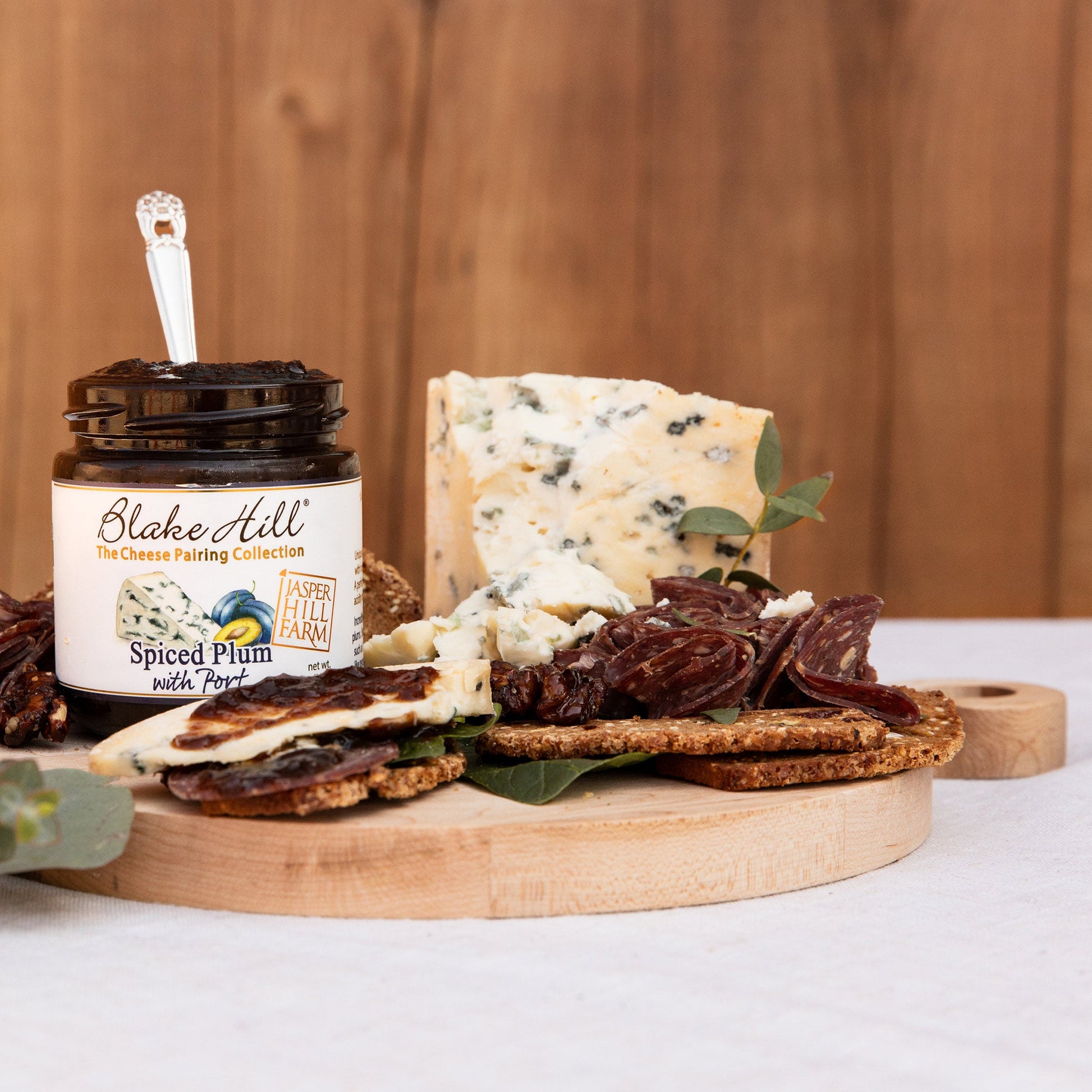 Platter of assorted cheeses, meats, and crackers with a jar of Blake Hill Spiced Plum Spread on a wooden board.