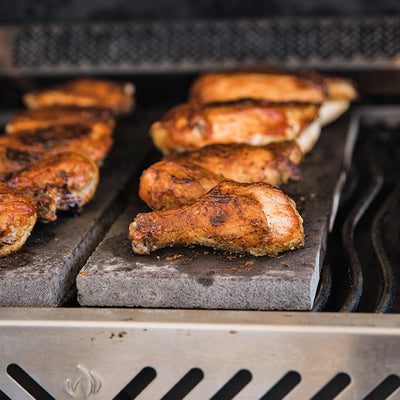 Grilled chicken drumsticks and thighs seasoned with spices are sizzling on a hot grill. The juicy chicken pieces are placed on a flat grilling stone, with a smoky background hinting at the outdoor cooking setting.