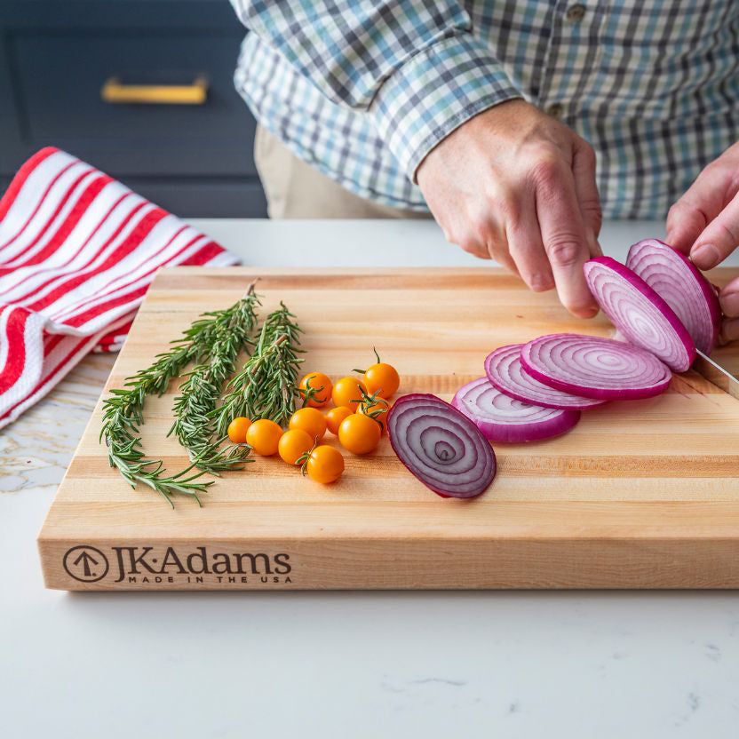 Person slicing red onions on a wooden cutting board with herbs and small fruits, branded 'JK Adams'.