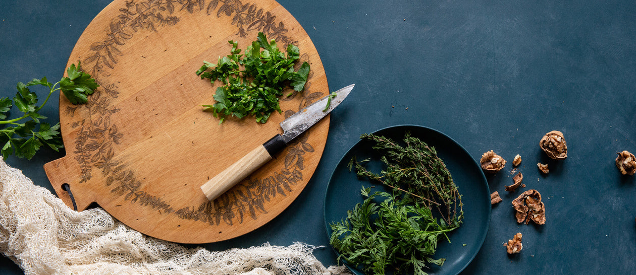 A styled flat-lay of a round wooden cutting board with engraved leaf patterns, featuring a small knife with a light wood handle and chopped green herbs, alongside a dark plate holding fresh herbs and scattered walnut shells on a dark blue surface.