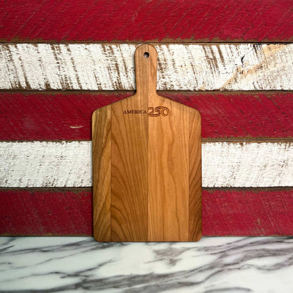Cherry serving board with the American 250 logo against a red and white stripes.

