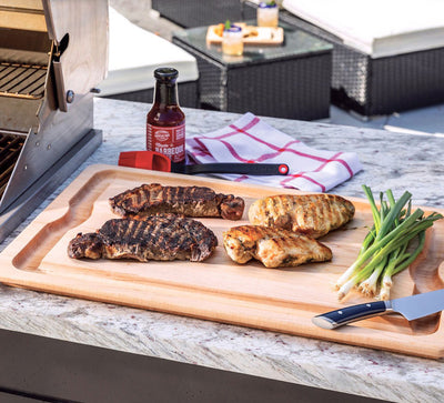 Grilled meats on a cutting board with a bottle of sauce and vegetables outdoors.