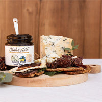A jar of Blake Hill Spiced Plum cheese with food styling including blue cheese, crackers, and dates on a wooden board against a wood panel background.
