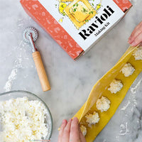 A top-down view of a ravioli making kit on a light marble surface, showing the product box, a wooden-handled rolling pin, a bowl of white dough, and hands placing stuffed pasta shapes onto a sheet of yellow pasta.