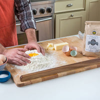 A person in an orange apron mixes flour with a cracked egg on a wooden cutting board in a kitchen, with butter, eggs, and a bag of Millhouse Organic Flour nearby.