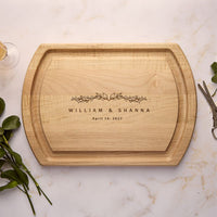 A personalized wooden serving board with rounded corners, featuring engraved text reading 'WILLIAM & SHANNA April 16, 2023' with a small floral motif. The board is photographed from above on a light marble surface with decorative greenery and a glass rim at the edges.