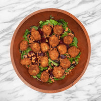 A top-down view of a wooden bowl filled with a salad featuring golden-brown fried balls, green spinach, mixed greens, and crushed peanuts, set on a white marble surface.