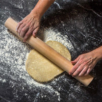 A wooden rolling pin being used by hands to roll out a round piece of dough on a dark, flour-dusted surface.
