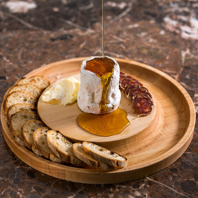 A wooden serving board on a dark marble surface holds a cheese dip or brie-like cheese in a small container with honey dripping into it, alongside sliced bread, butter, and cured sausage.