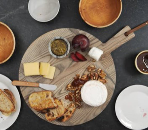 A wooden board with assorted cheeses, sliced bread, walnuts, and a small bowl of pesto. Two circular wooden bowls and two white plates are placed around the board on a dark surface.