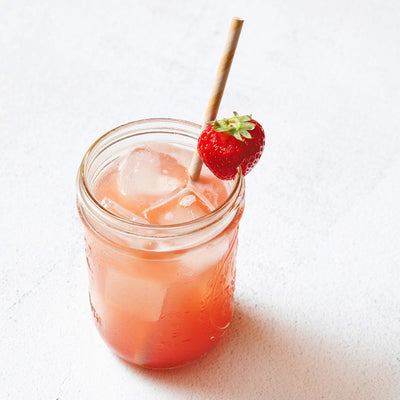 A refreshing pink drink in a mason jar with ice cubes, garnished with a strawberry on the rim and a striped paper straw. The background is a textured white surface.
